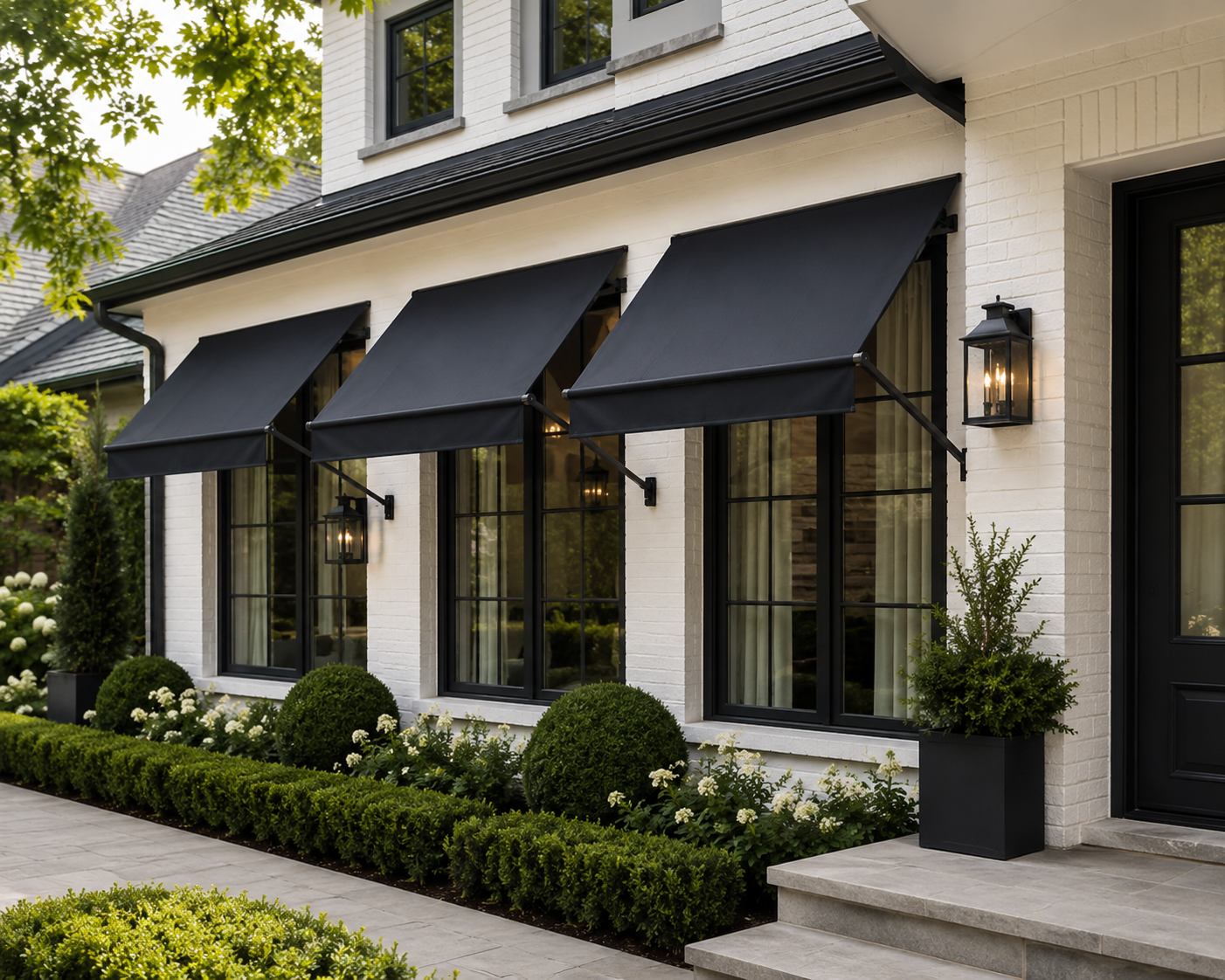 Exterior of a modern house with three large windows with black frames and black awnings, white brick walls, potted plants, and landscaped shrubs.