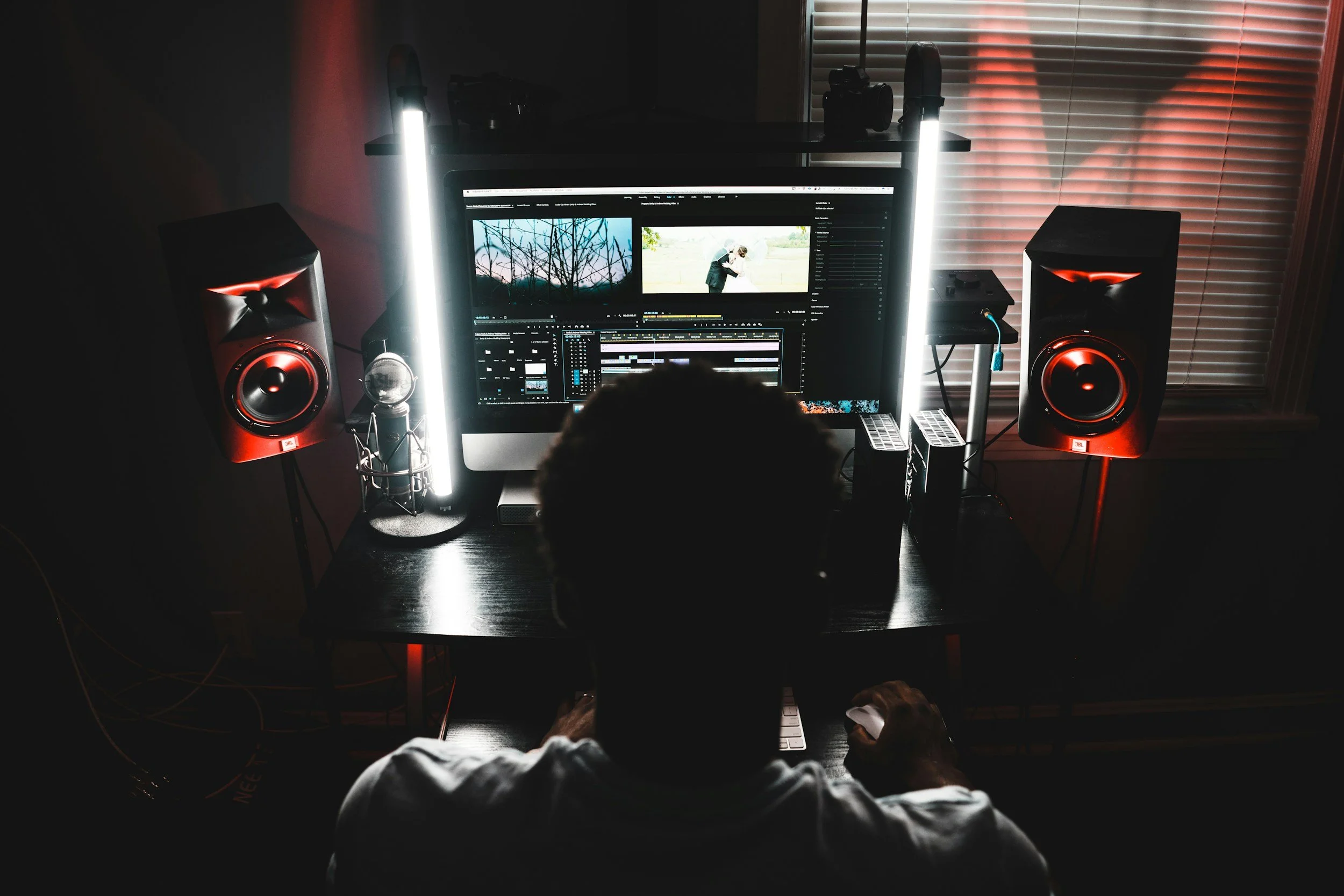 A dimly lit workspace with a person working on a computer with dual monitors, surrounded by studio speakers, a microphone, and studio lights, with window blinds in the background.