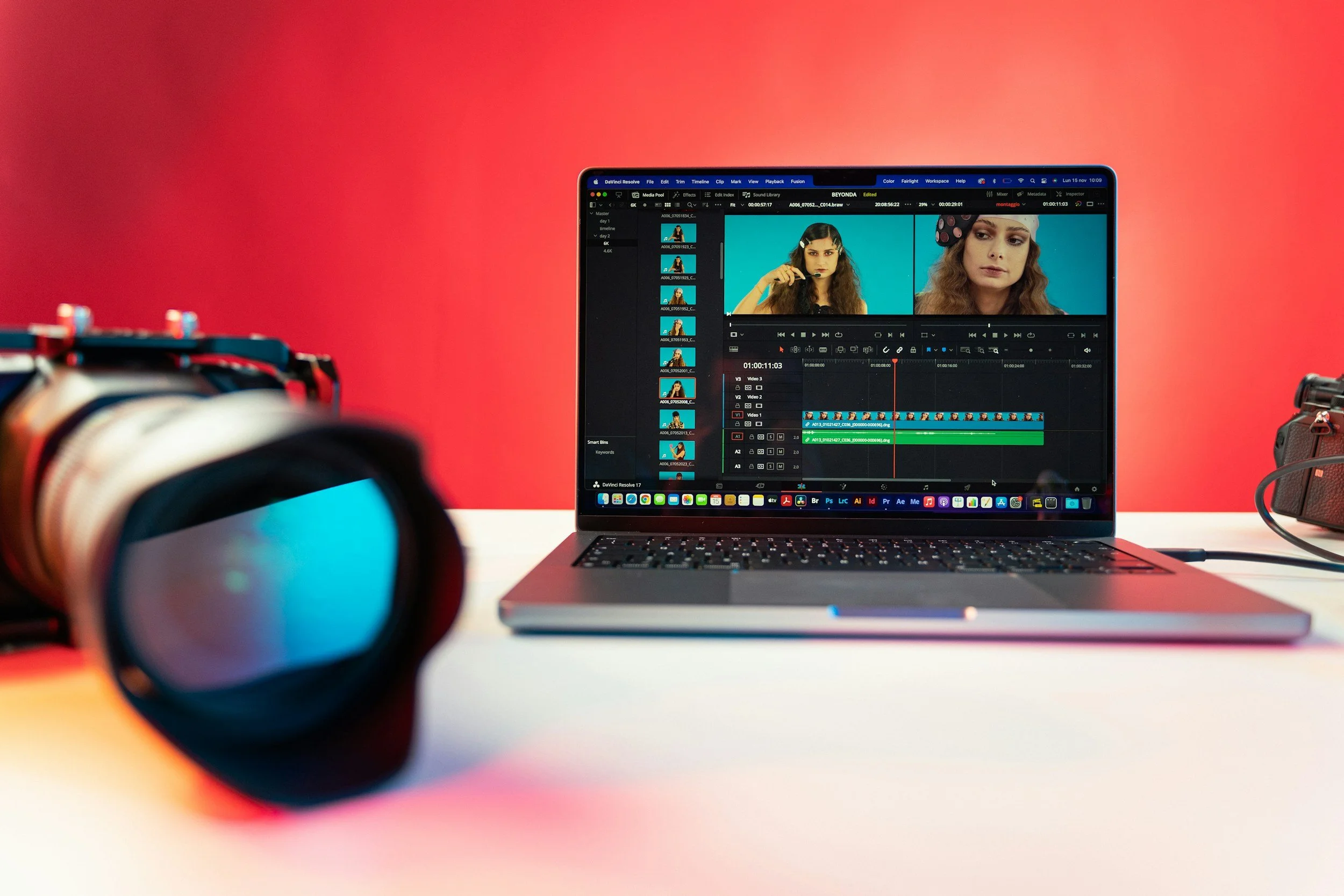 A laptop displaying video editing software with two frames of a woman with curly hair and a headband, on a white table with a blurred camera in the foreground and a vibrant red background.