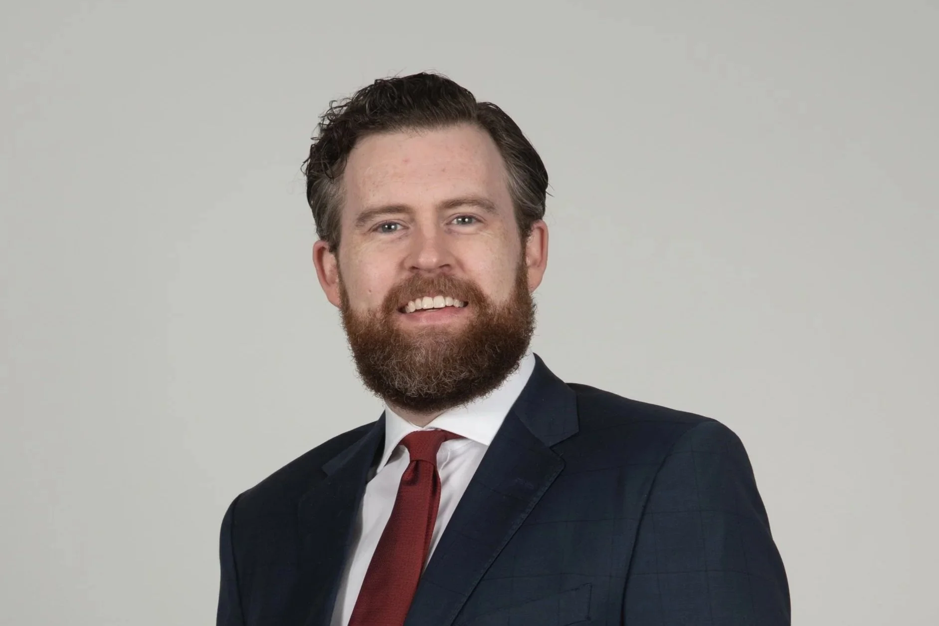 Portrait of a man with a beard, smiling, wearing a dark suit, white shirt, red tie, and standing against a plain light gray background.