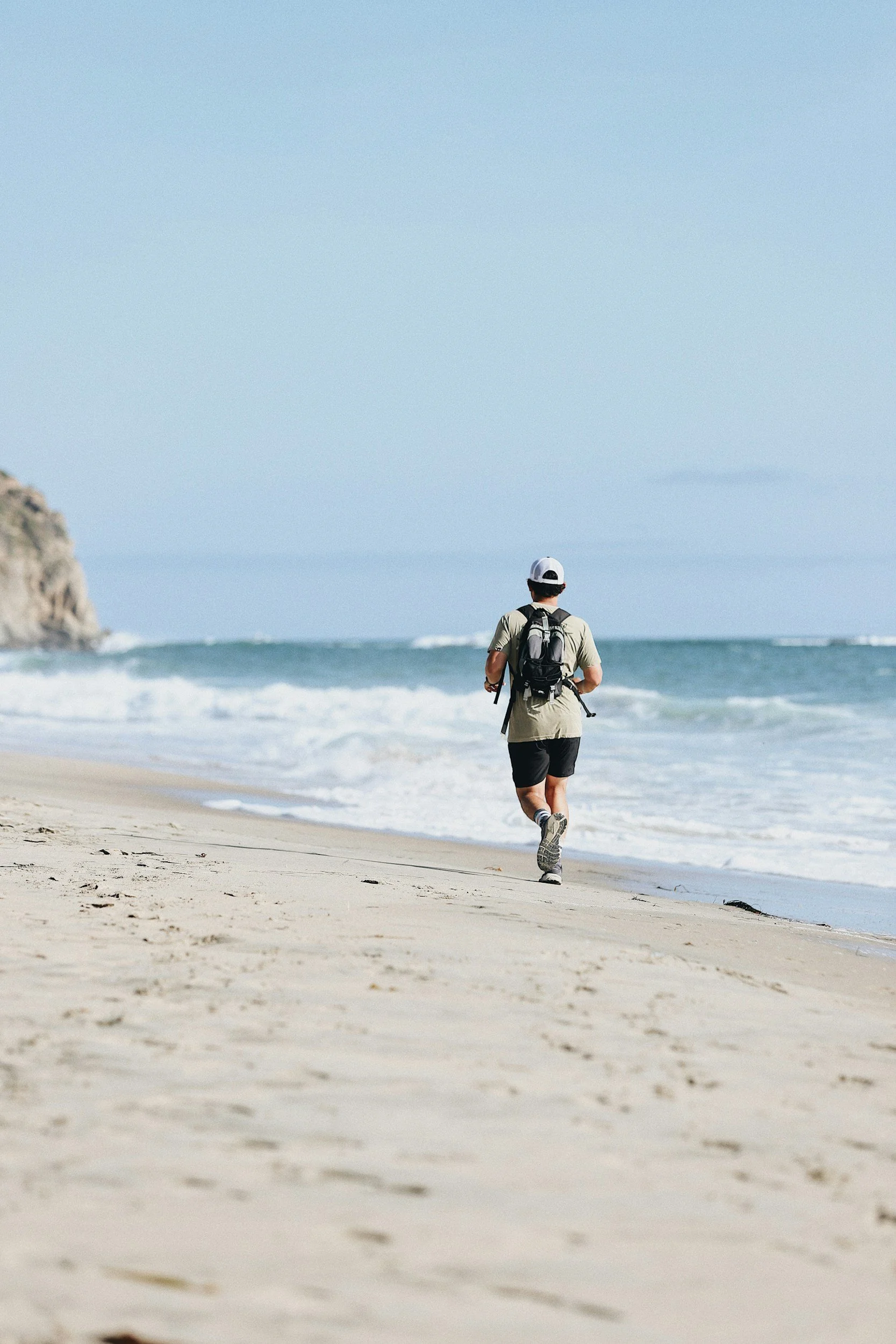 A man walks along the beach with waves crashing onto the shore, wearing a white cap, a backpack, and sporty clothing on a sunny day.