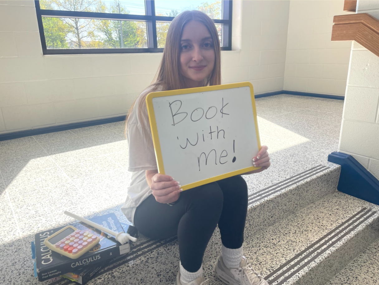 A young woman with long hair sitting on a staircase holds a whiteboard that says, 'Book with me!' She is inside a building near a window with trees outside, wearing a white shirt, black pants, and white sneakers. There are books and a calculator on the steps beside her.