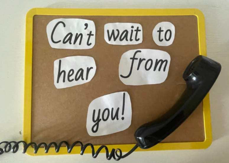 A corkboard with cut-out paper pieces spelling "Can't wait to hear from you!" and a black landline telephone resting on the bottom right corner.