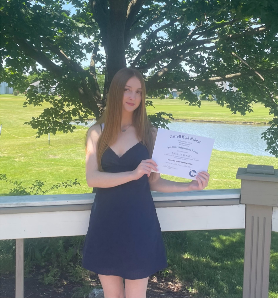 A young woman with long hair wearing a dark dress holding a high school diploma standing outdoors in front of a tree and a pond, celebrating her academic achievement.