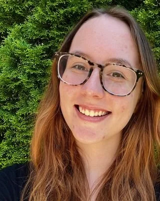 A young woman with long reddish-brown hair and glasses smiling in front of green foliage.