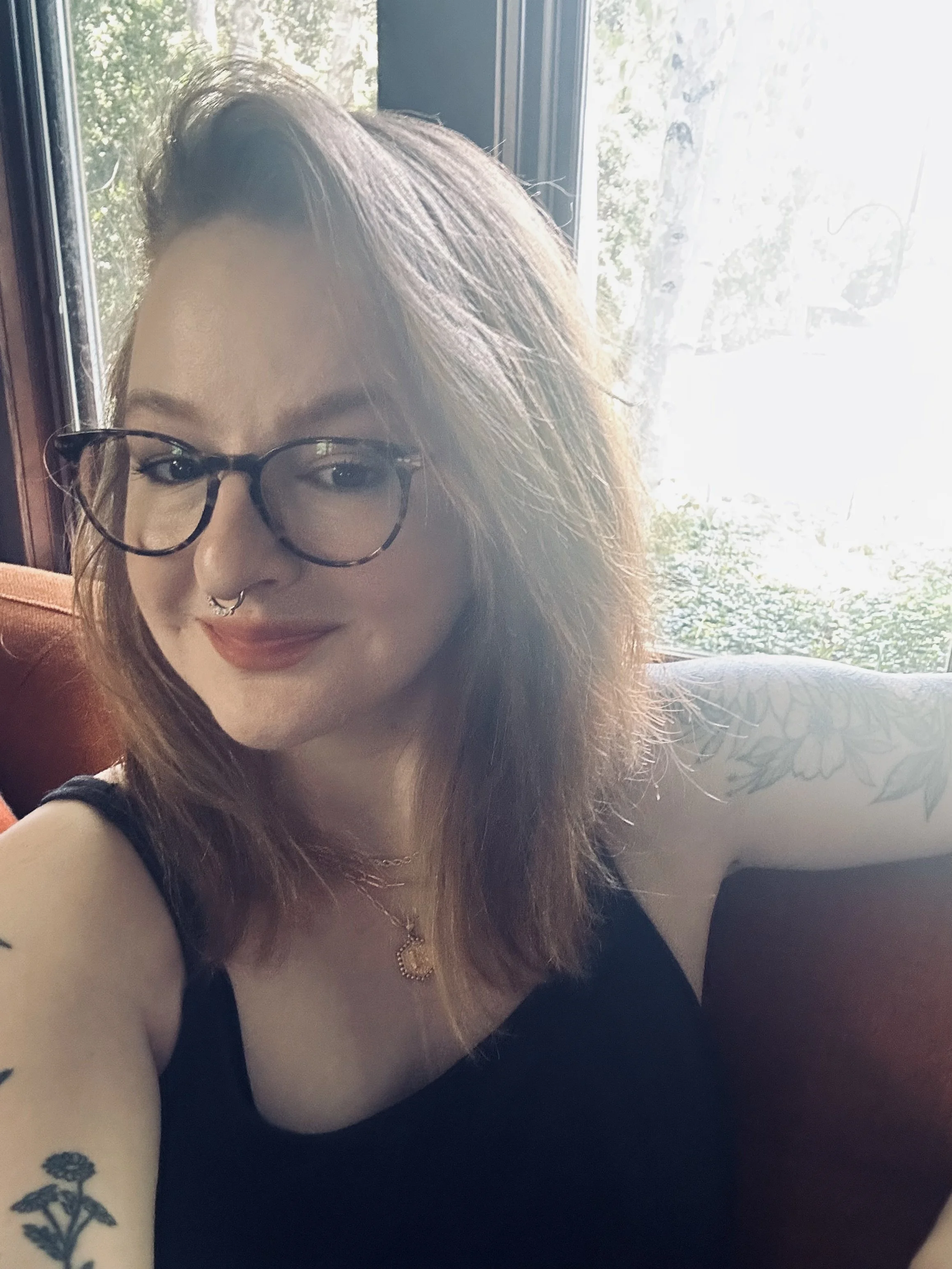 A woman with glasses and septum piercing smiling, with tattoos on her arm, sitting indoors near a window with trees outside.