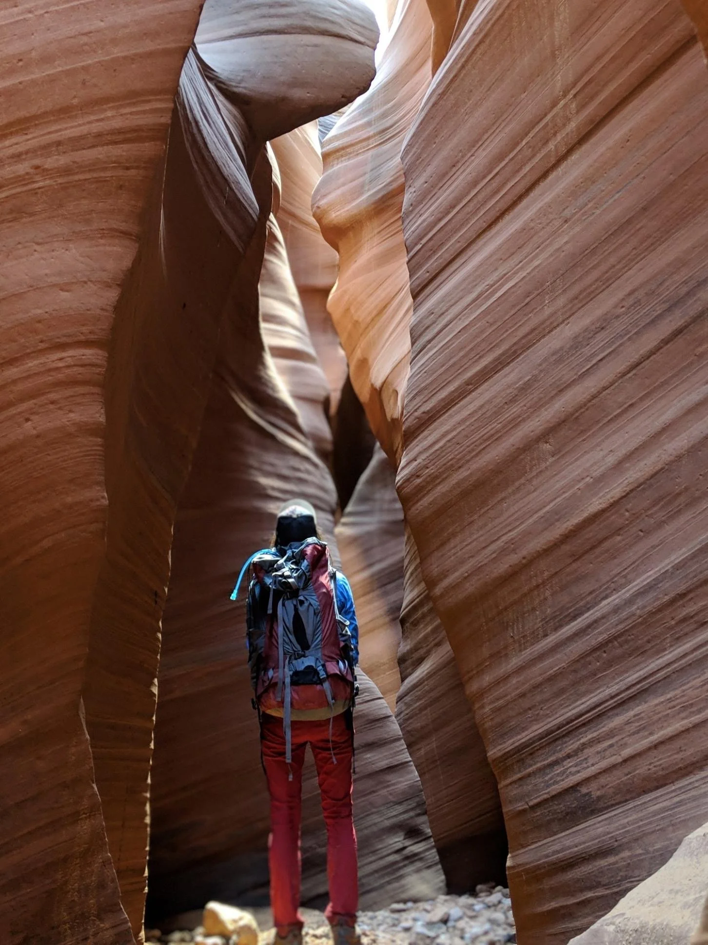 A person wearing a backpack and hiking gear exploring a narrow sandstone canyon with smooth, curved rock walls.