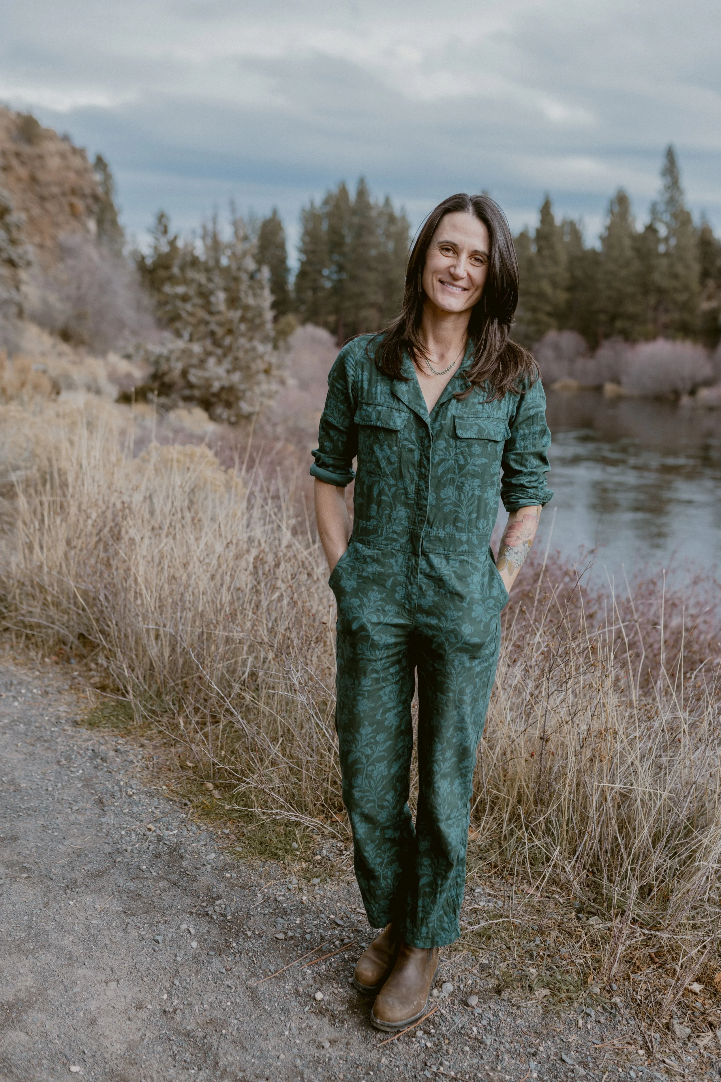 A woman standing outdoors near a river and trees, smiling, wearing a green patterned jumpsuit, brown boots, with one hand in her pocket.