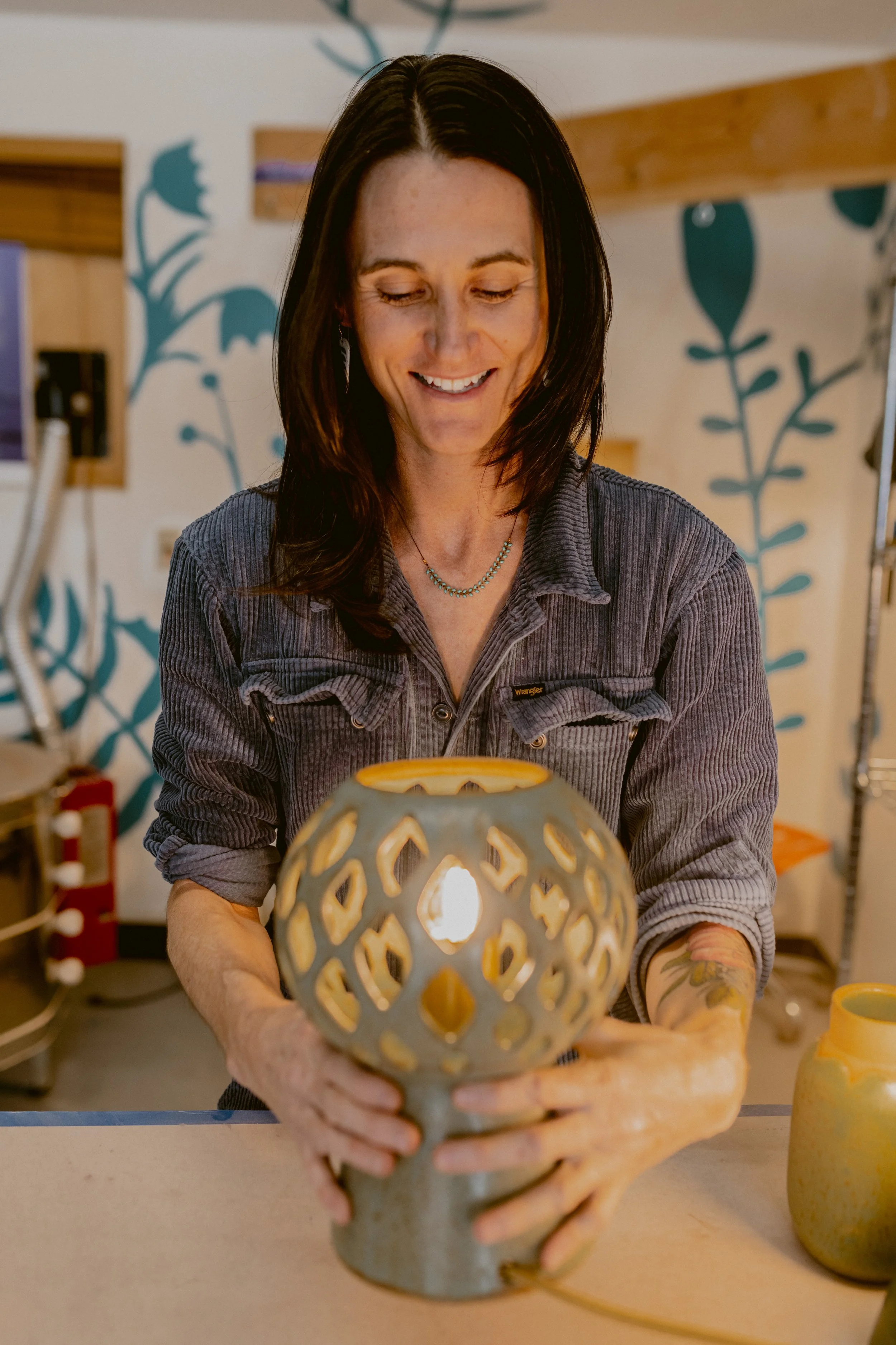 Woman with dark hair and a necklace holding a decorative ceramic lamp with a light inside, in a room with painted wall art and other pottery on the table.