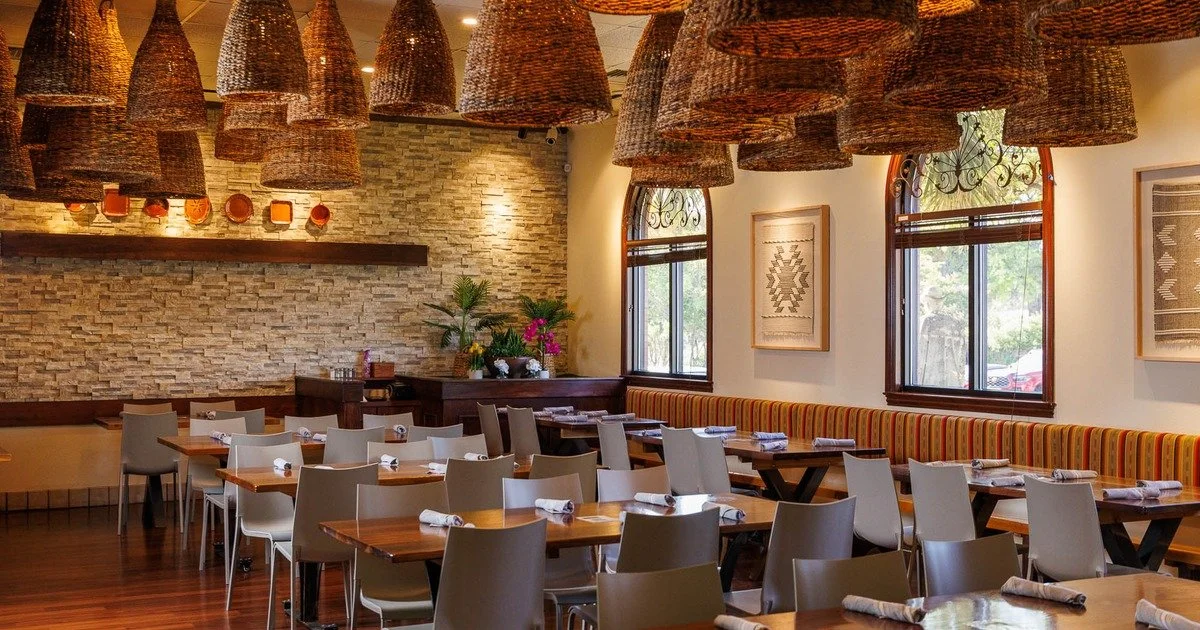 Interior of a restaurant with wooden tables and chairs, decorated with woven pendant lights hanging from the ceiling, stone accent wall, framed artwork, and windows with view outside.