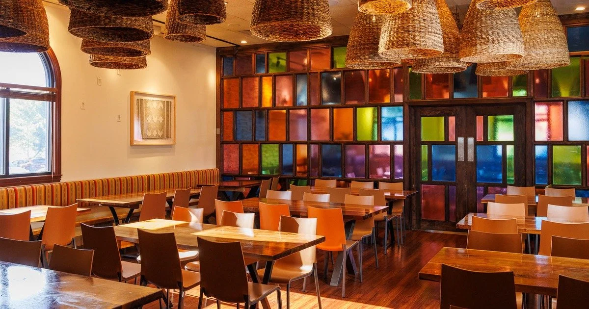 Interior of a restaurant with wooden tables and colorful chairs, decorated with a multicolored stained glass wall and woven pendant light fixtures.