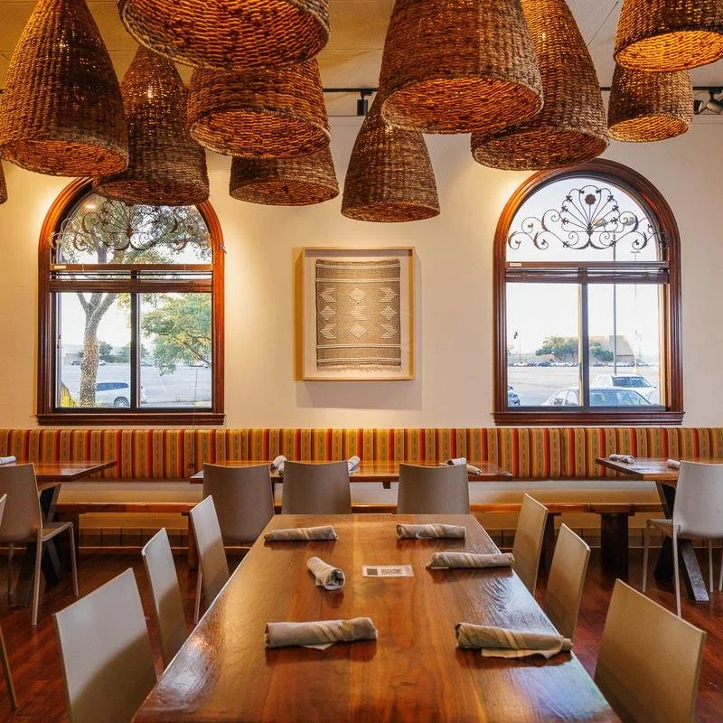 Interior of a restaurant with a wooden table set with napkins, a long striped upholstered banquette along the wall, three arched windows with decorative ironwork, and woven pendant light fixtures hanging from the ceiling.
