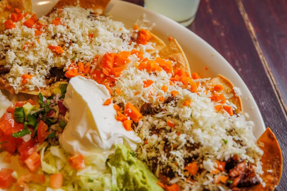 Close-up of a plate with Mexican food, including tacos topped with cheese, mayonnaise, and chopped carrots, along with diced tomatoes, lettuce, and sour cream.