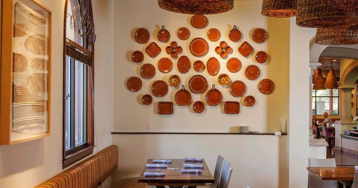 Interior of a restaurant with a wall decorated with various terracotta plates and bowls, a window with wooden blinds, and a dining table set with napkins.