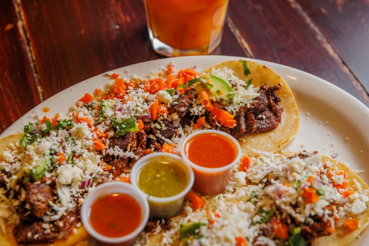 Three tacos on a white plate with assorted toppings and three salsa dips, with a glass of iced tea in the background.