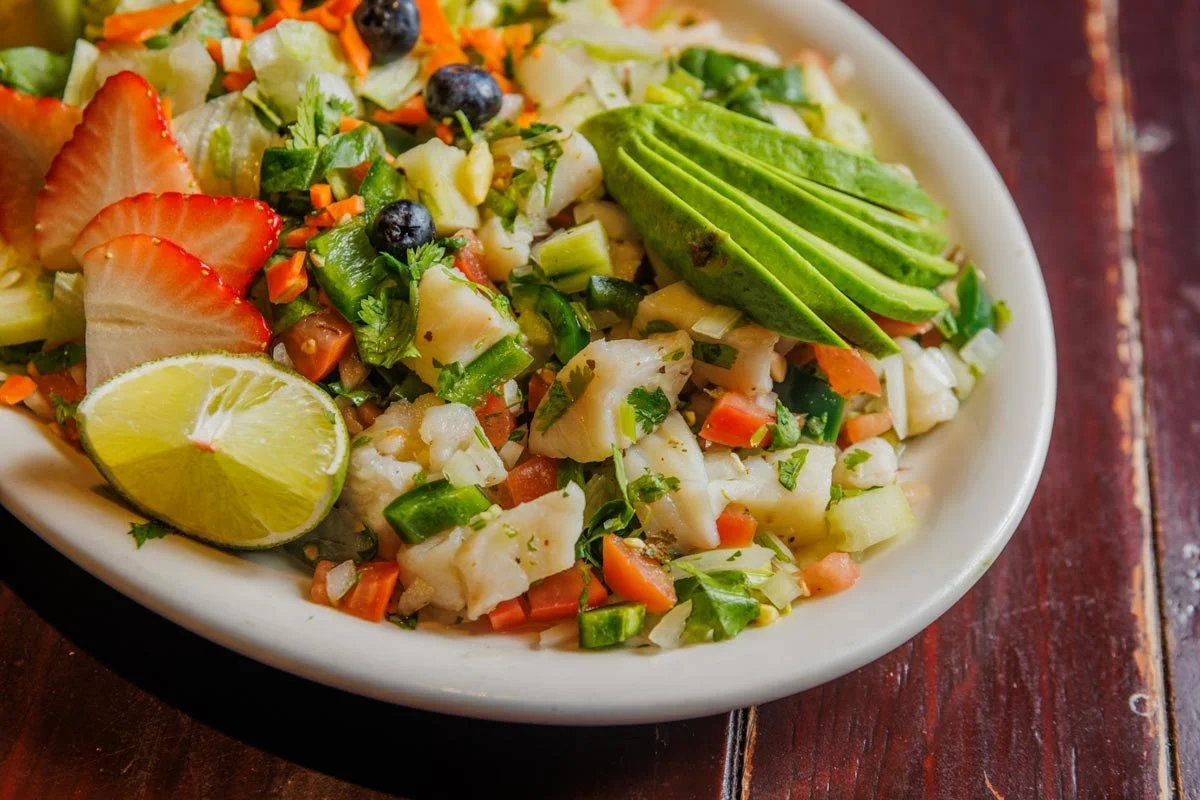 A bowl of seafood ceviche garnished with sliced avocado and lime, containing chopped vegetables, fruit, and herbs.
