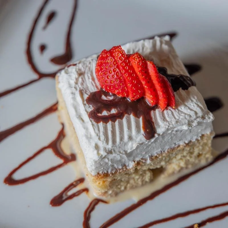 Slice of vanilla cake with whipped cream, topped with strawberries and chocolate, drizzled with chocolate syrup, on a white plate with chocolate syrup design.