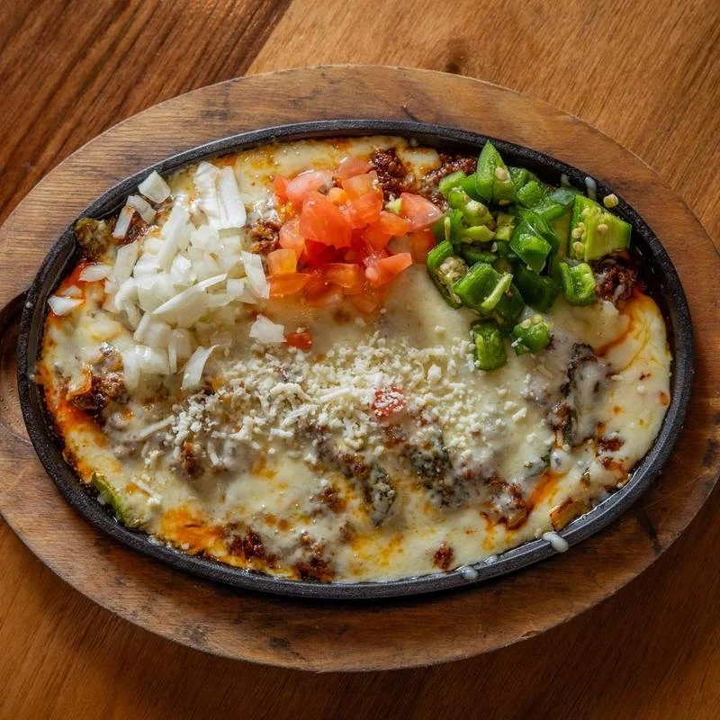 Mexican-style cheese enchilada casserole topped with shredded cheese, diced tomatoes, chopped onions, and sliced green onions served on a hot cast iron skillet on a wooden serving board.