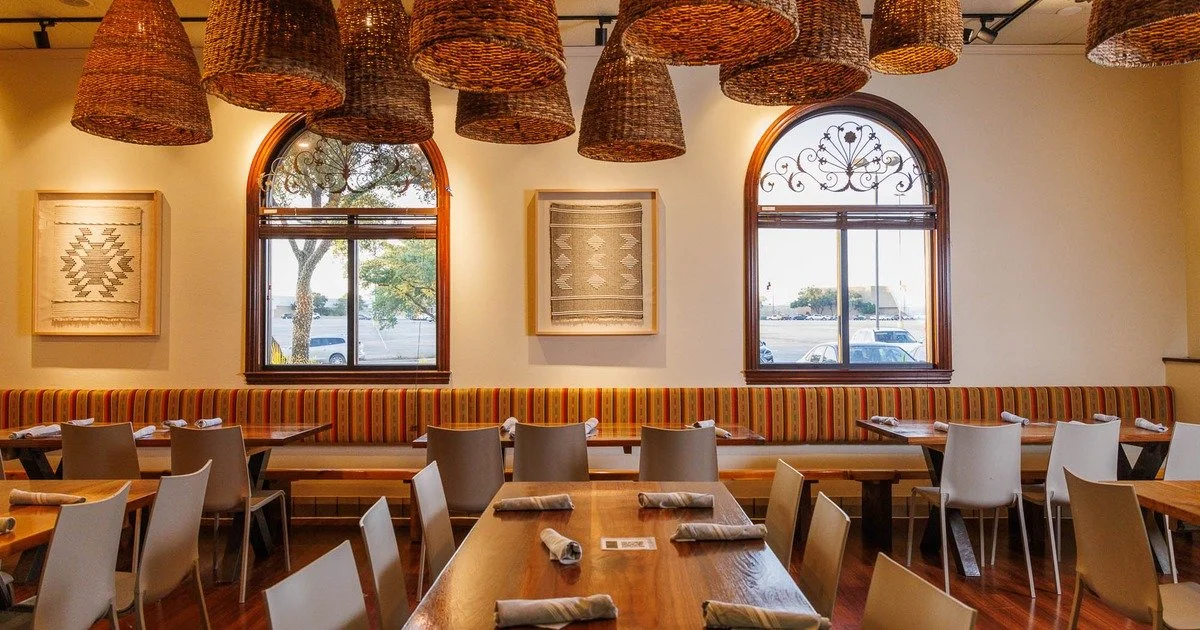 Interior of a restaurant with wooden tables and white chairs, colorful striped banquette seating, woven pendant lights, and two arched windows with decorative ironwork, overlooking parked cars and trees outside.
