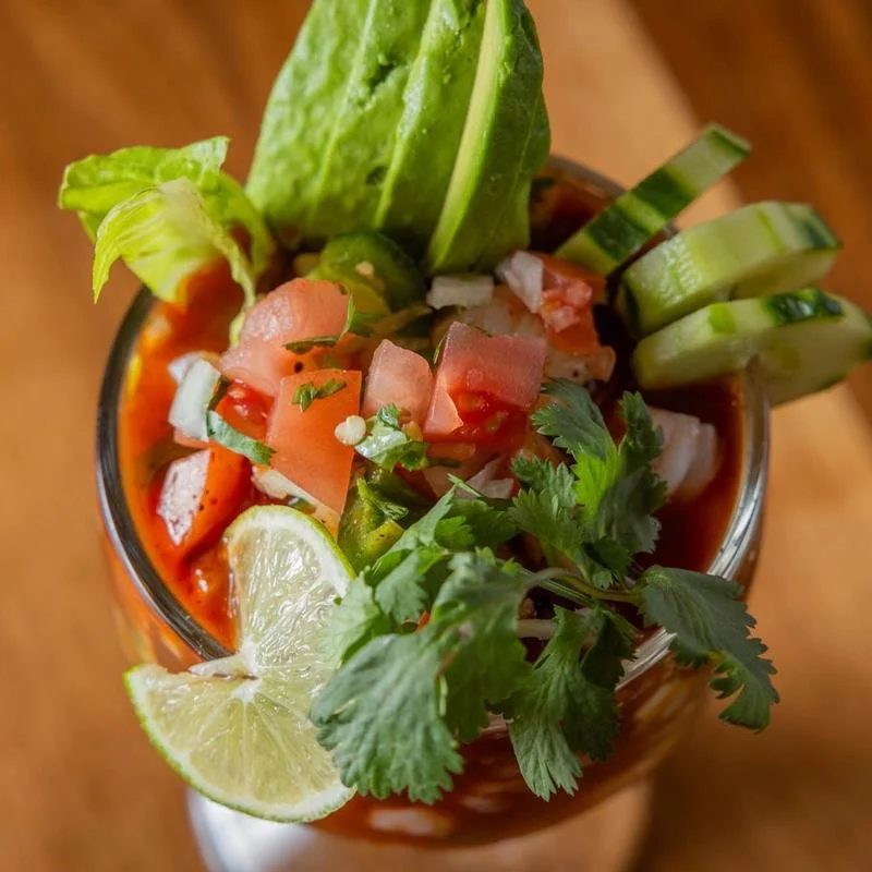 A bowl of tomato-based soup topped with chopped tomatoes, cucumbers, cilantro, lime wedge, lettuce, and possibly other fresh vegetables