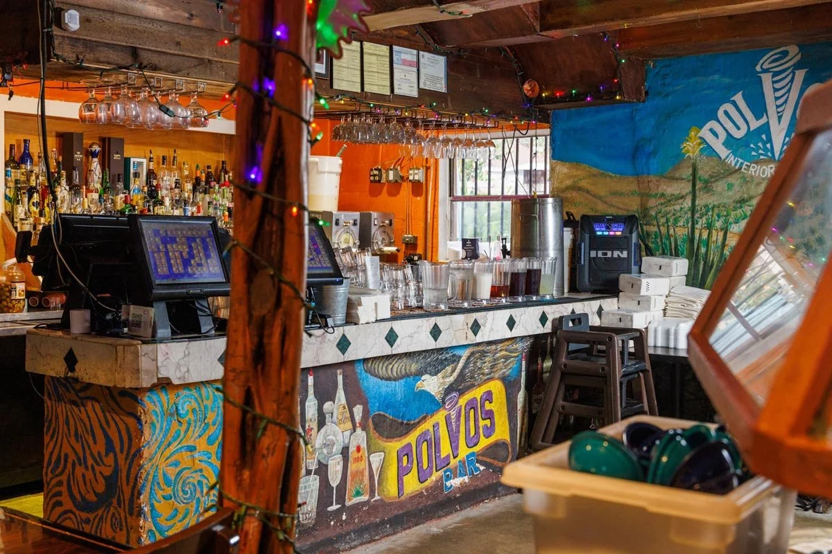 Interior of Pollos Bar with a colorful mural, bar counter with various bottles and glasses, cash registers, and drink dispensers. Bar stools are stacked, and colorful string lights decorate the wood-paneled ceiling and supporting post.