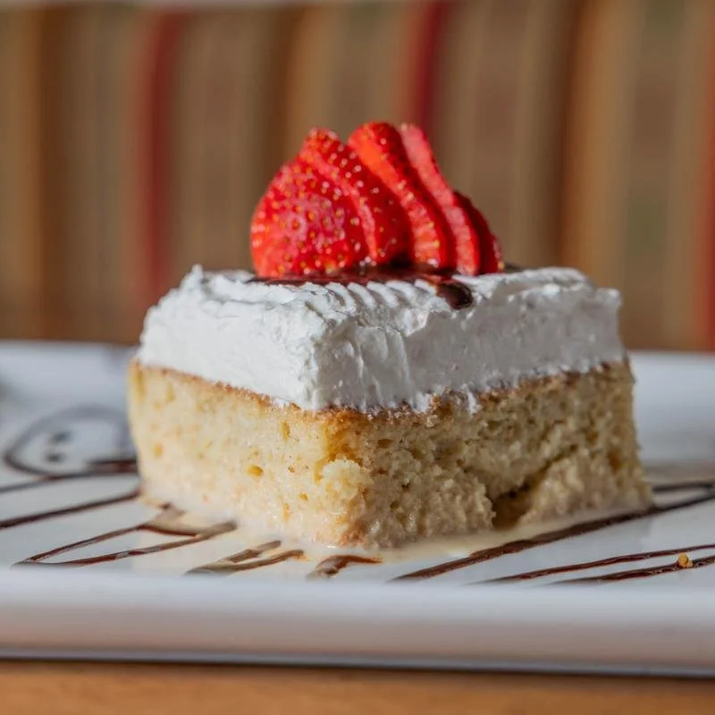 A slice of cake with white frosting, topped with sliced strawberries, served on a white plate with chocolate drizzle.
