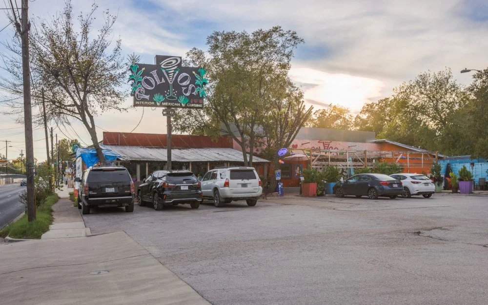 Parking lot in front of a mexican restaurant called "Ole" with cars parked, trees, and a sunset sky.
