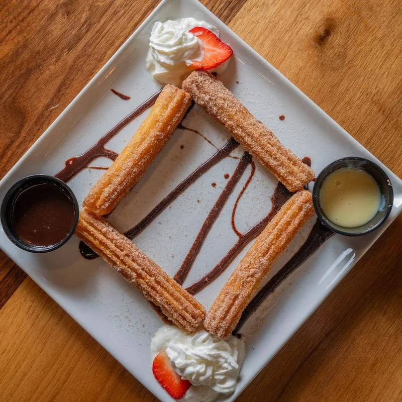 A dessert plate featuring four churros arranged in a square, garnished with whipped cream and strawberry slices, drizzled with chocolate and caramel sauces on a white square plate.