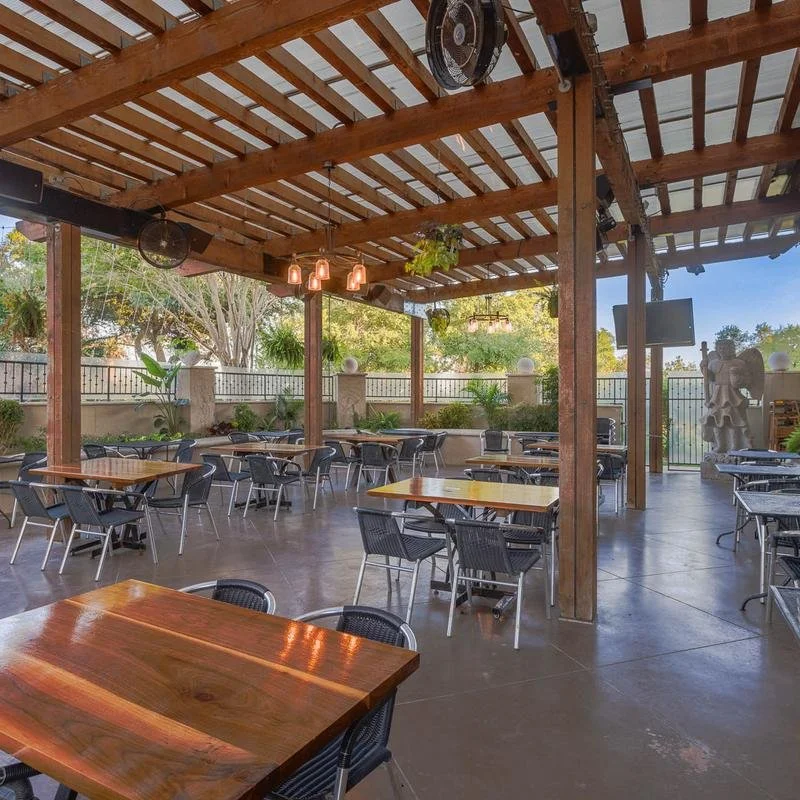 Outdoor patio with wooden roof, chairs and tables, potted plants, and a fence. Some hanging plants and a statue are visible.