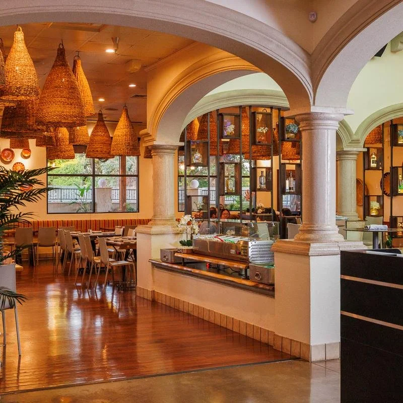 Interior of a restaurant with wooden tables, beige chairs, decorative wicker pendant lights, and large windows. The space features arches, columns, and decorative shelving with vases and plants.