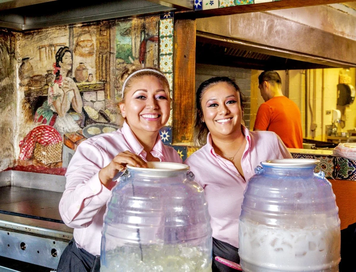 Two women in pink shirts smiling behind large drink dispensers in a restaurant with a colorful mural on the wall.