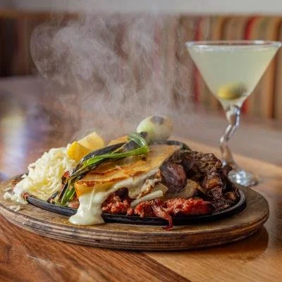 A sizzling plate of food with steam rising, including grilled meat, vegetables, and melted cheese, with a lemon wedge and a drink in a martini glass in the background.
