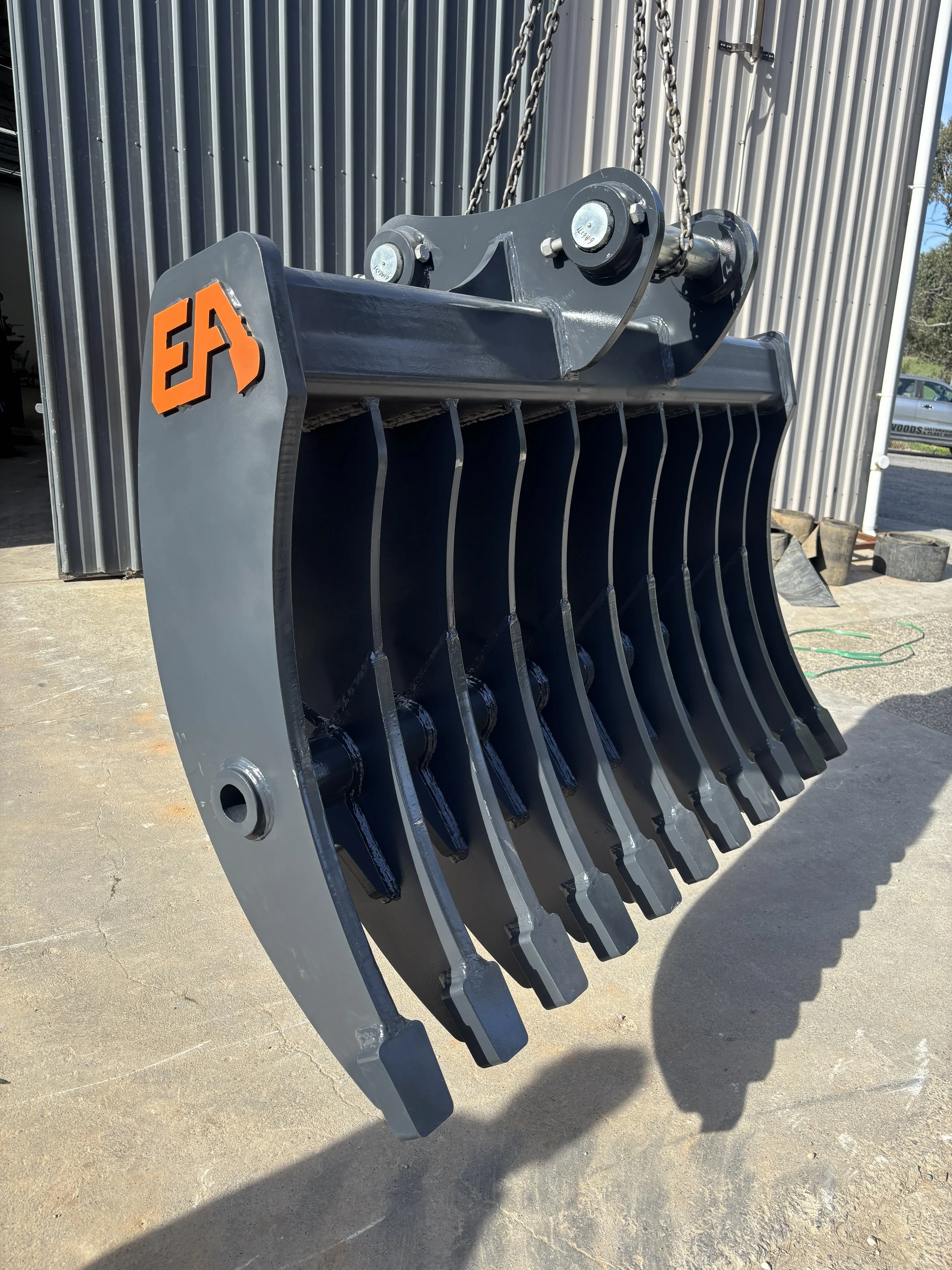 A large stick rake attachment with the logo 'EA' in orange, hanging on chains outside a building with vertical metal siding.