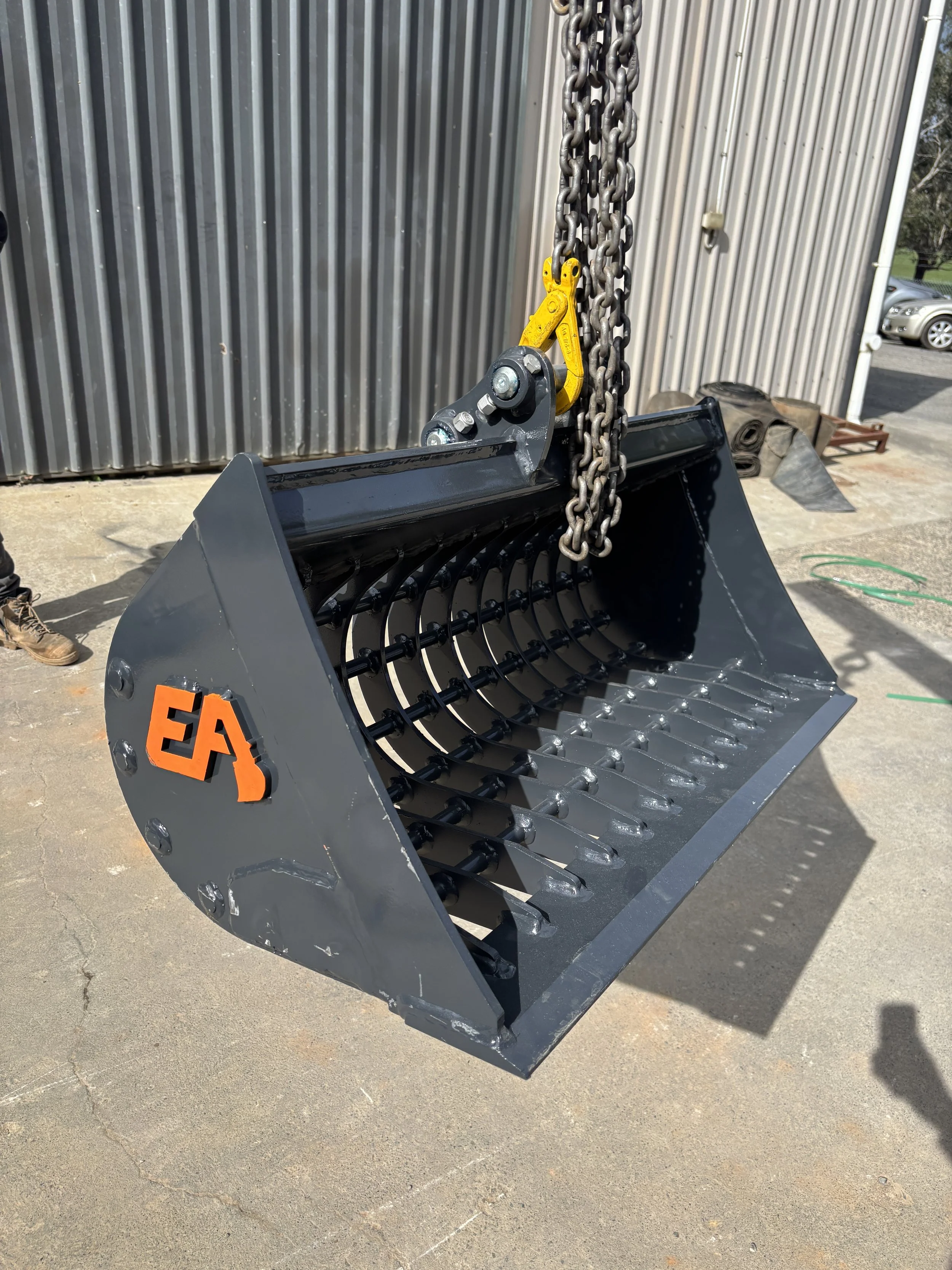 A sieve bucket for an excavator hanging from chains, and a yellow link, shown outdoors in front of a metal building.
