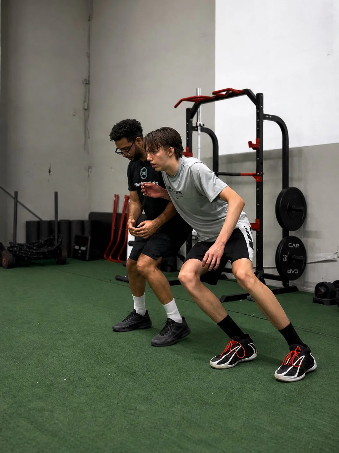 Two young men training in a gym. One is helping the other with a squat exercise, both are dressed in workout clothes. The gym has green turf flooring and workout equipment like weights and a pull-up rig in the background.