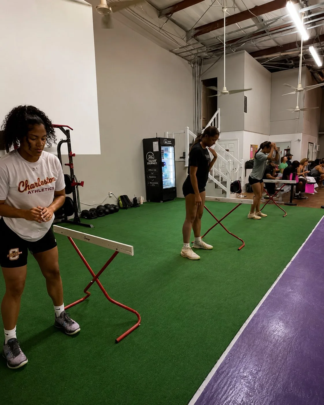 Three women are performing step-up exercises on red and white aerobic steps in a gym with green turf flooring. Other people are sitting at tables in the background, and a vending machine is visible against the wall.