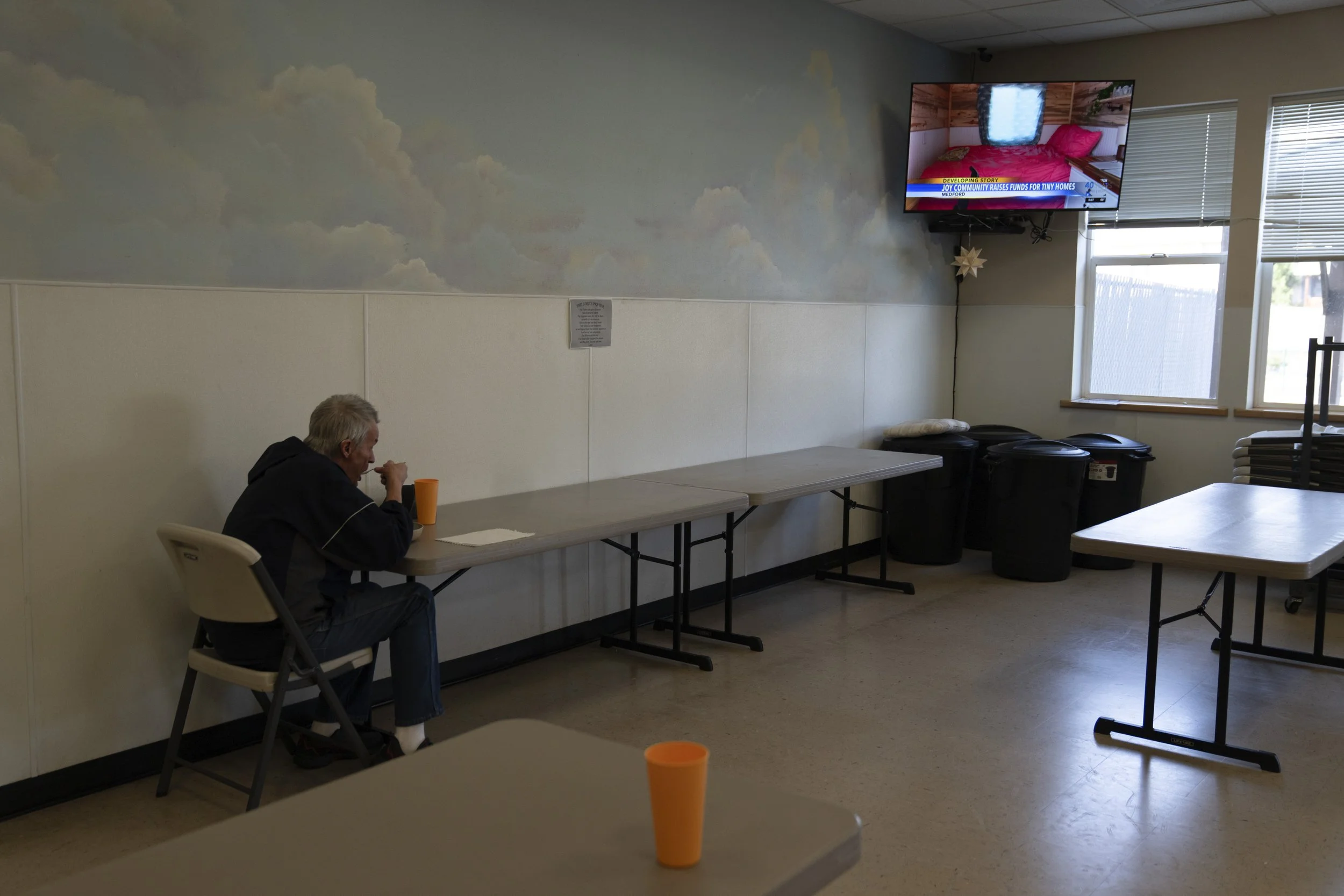 A resident at the Gospel Rescue Mission homeless shelter watches a report by KDRV NewsWatch 12 on the community raising money for tiny homes while eating dinner Thursday, March 21, 2024, in Grants Pass, Ore.