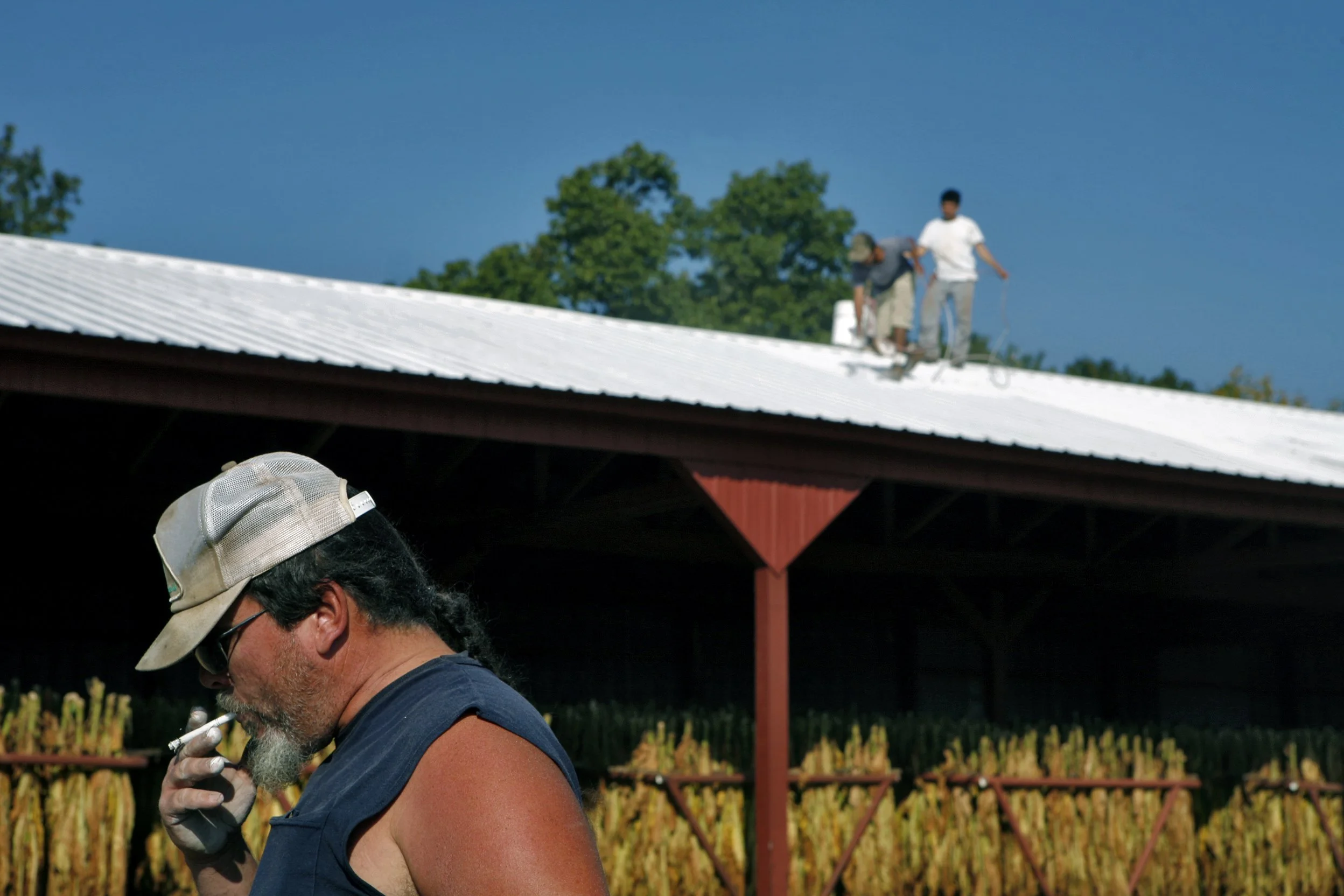 Chad Barber takes a smoke break while the migrant workers on his family farm, Cortes and Rodriguez, finish spray painting the top of the tobacco barn’s roof. 