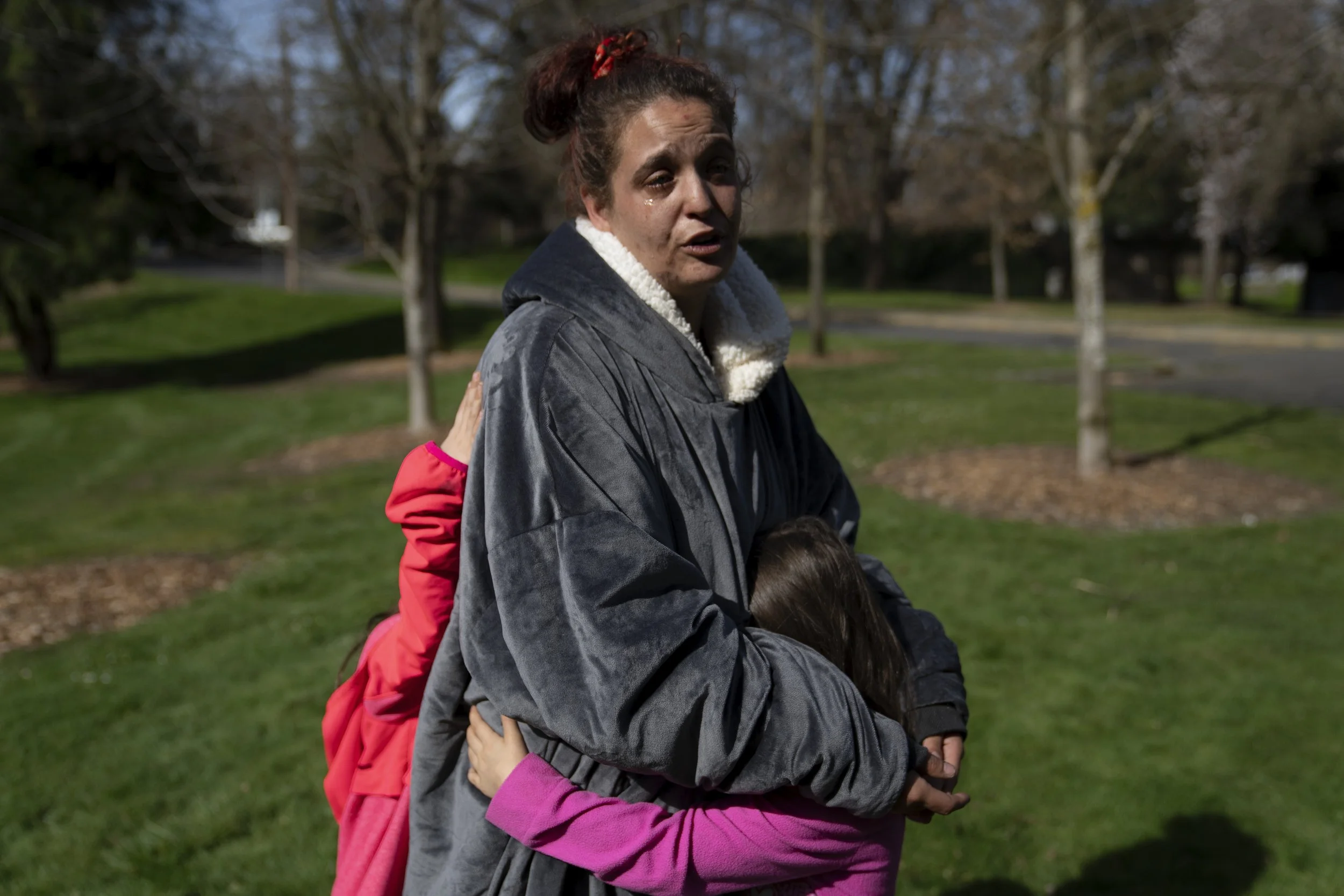 Nichole McKendrick holds her two daughters as she speaks after getting free food at Riverside Park on Thursday, March 21, 2024, in Grants Pass, Ore. McKendrick has been living out of a vehicle with her two girls. 