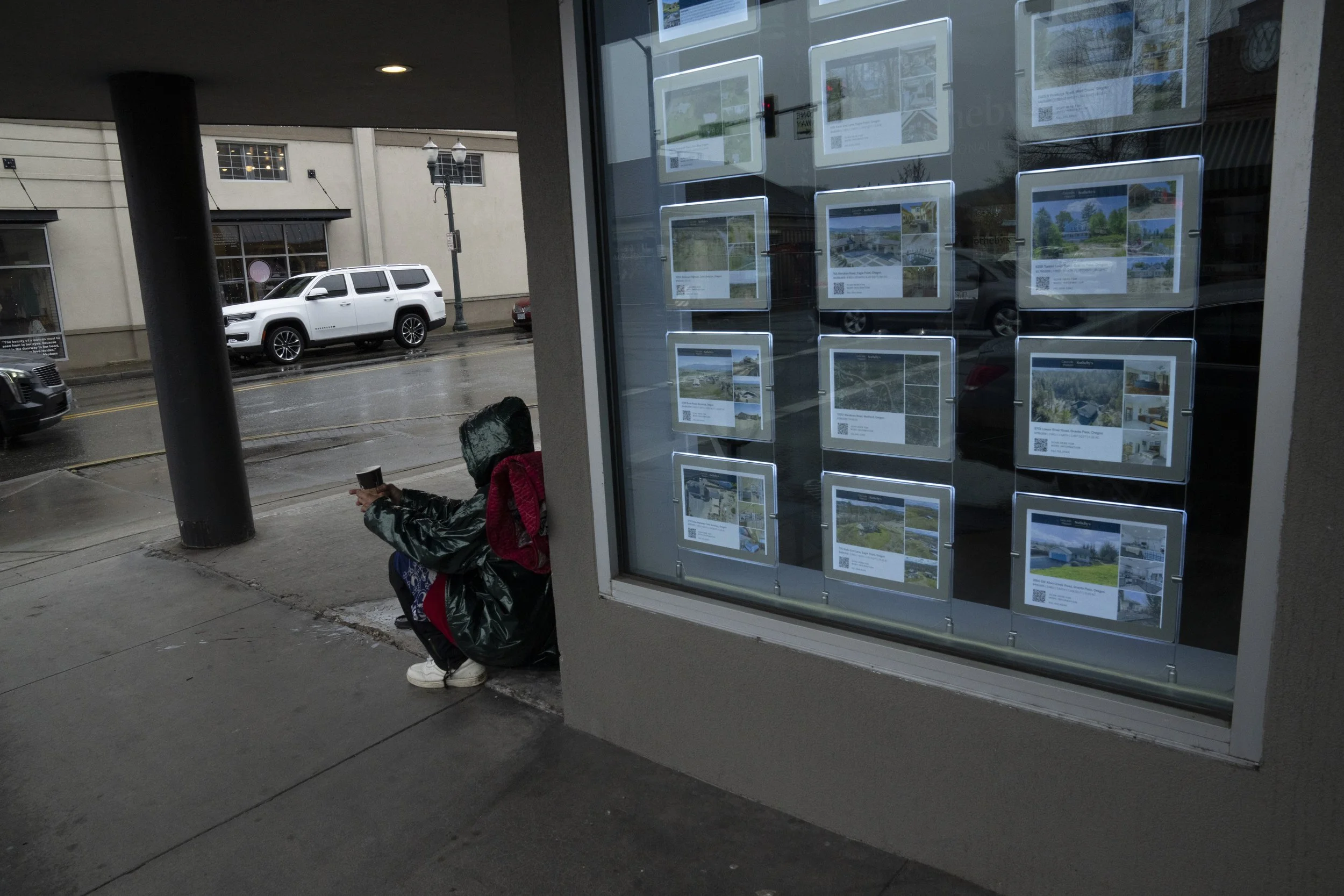 A homeless person sits next to a window displaying houses for sale on Saturday, March 23, 2024, in Grants Pass, Ore.