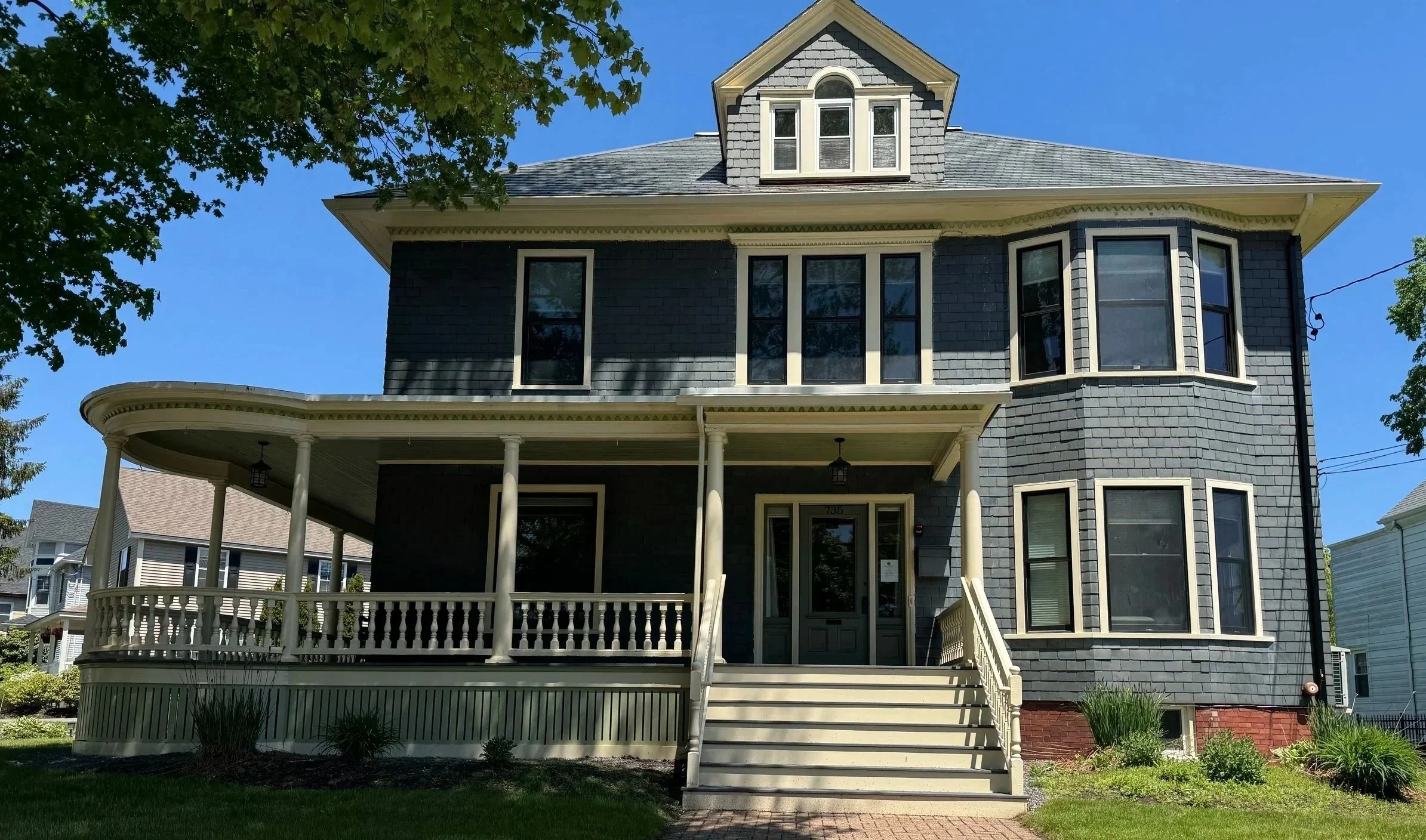 A multi-story house with a curved front porch supported by white columns, a small set of stairs leading to the front door, gray shingles, and a large bay window. There is a tree with green leaves partially visible on the left side and a clear blue sky.