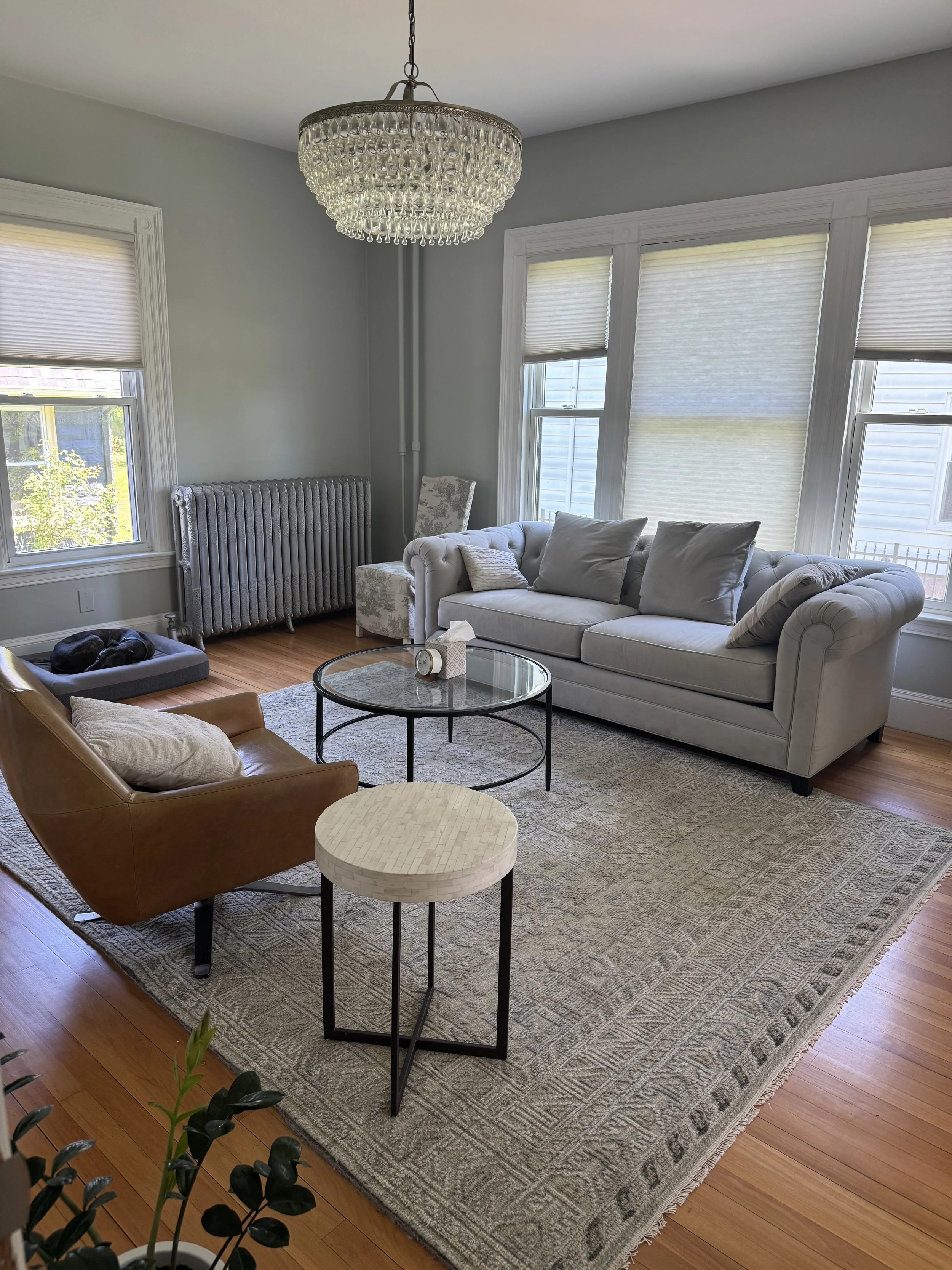 Living room with light gray walls, white window trim, large window with blinds, a beige tufted sofa with pillows, a brown armchair, a light-colored area rug, a glass coffee table, a patterned chair, a fireplace with a radiator, and a chandelier with crystal drops.