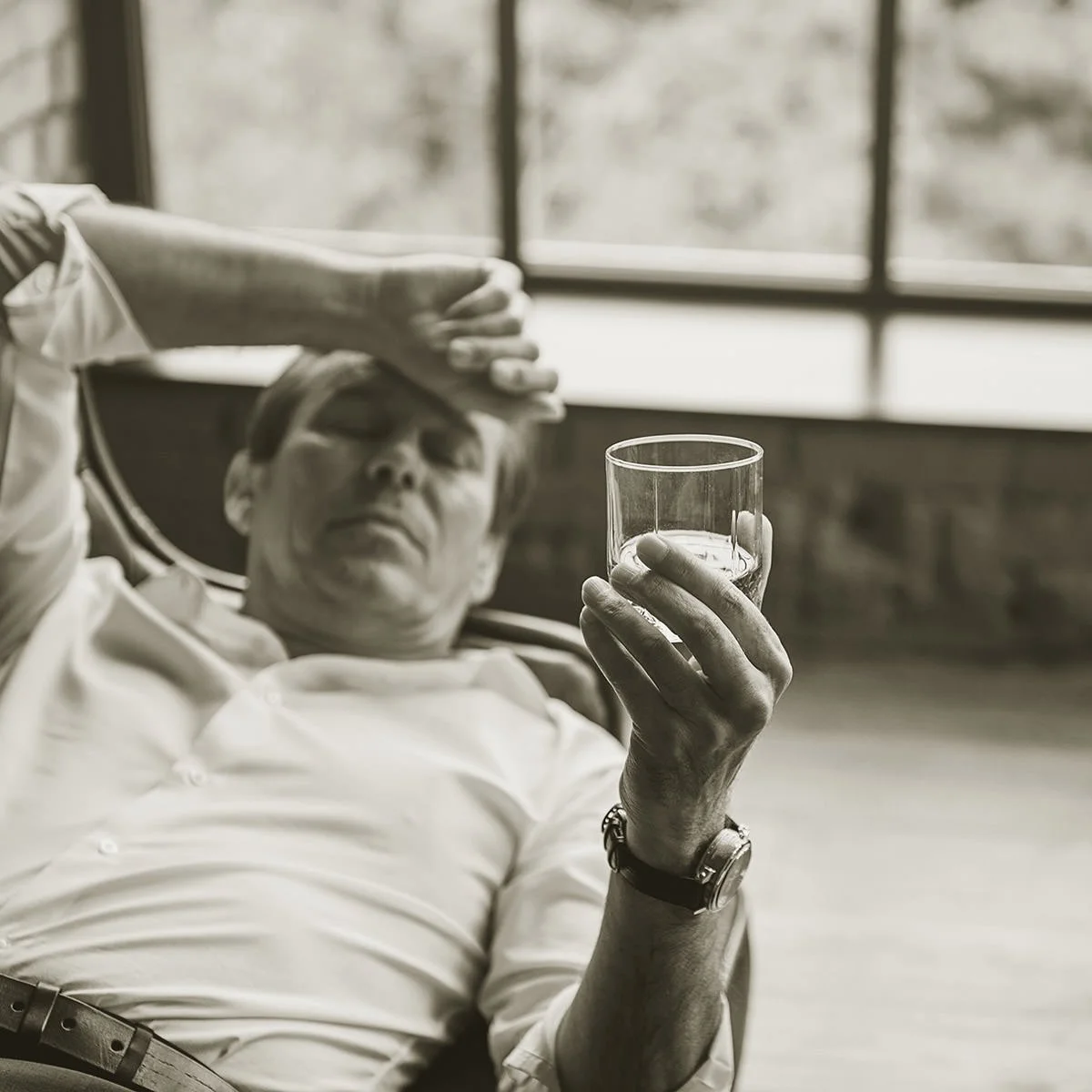 A man is reclining in a chair, holding a glass of whiskey, with his other hand resting on his forehead. The scene is in a room with large windows in the background, black and white photograph.