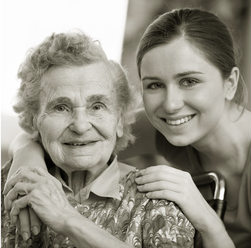 A young woman smiling and sitting close to an elderly woman, both with arms around each other, in a close-up portrait in black and white.