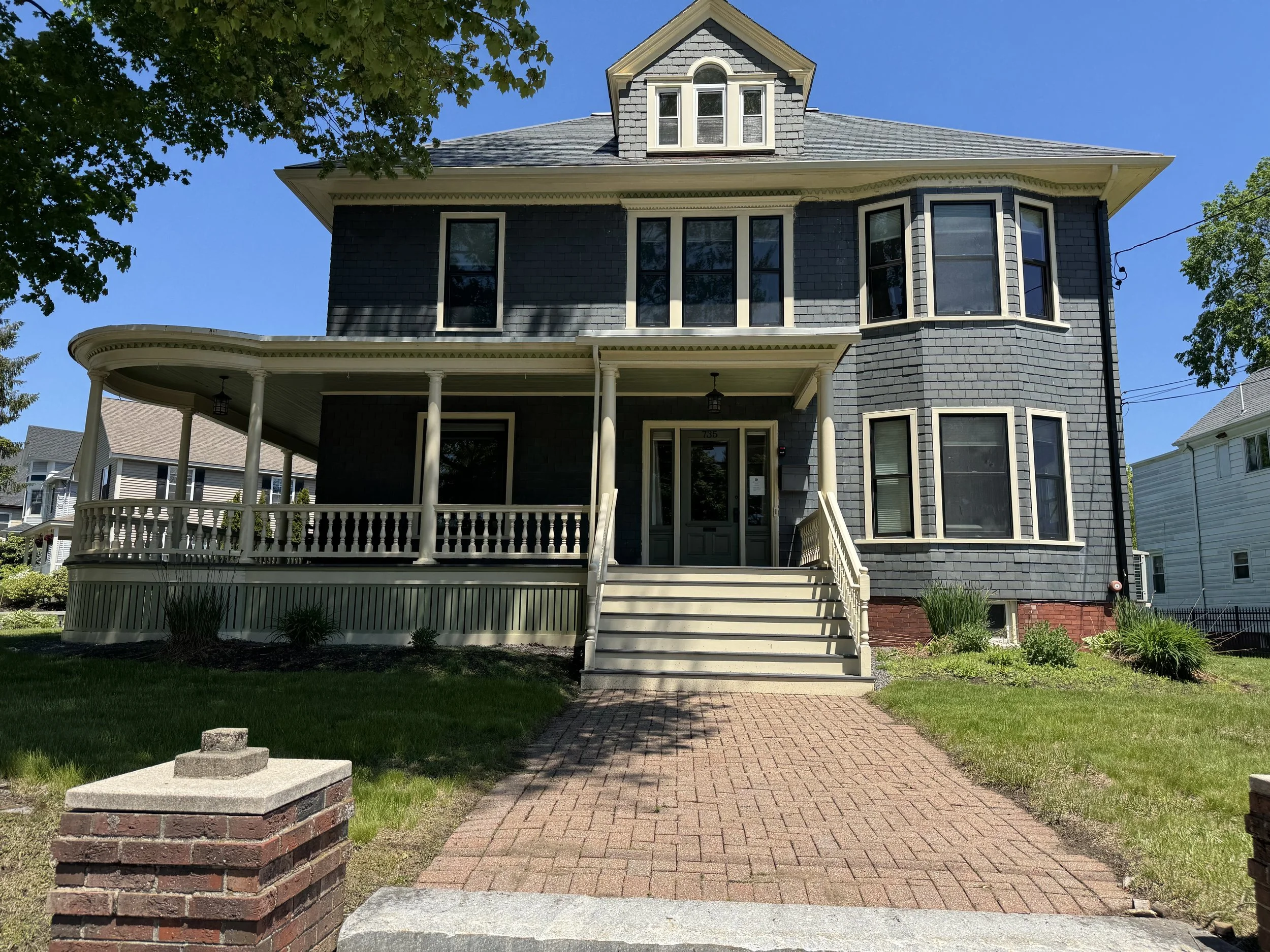 Front view of a gray, three-story house with a covered porch, surrounded by green grass, trees, and a brick pathway leading to the entrance, under a clear blue sky.