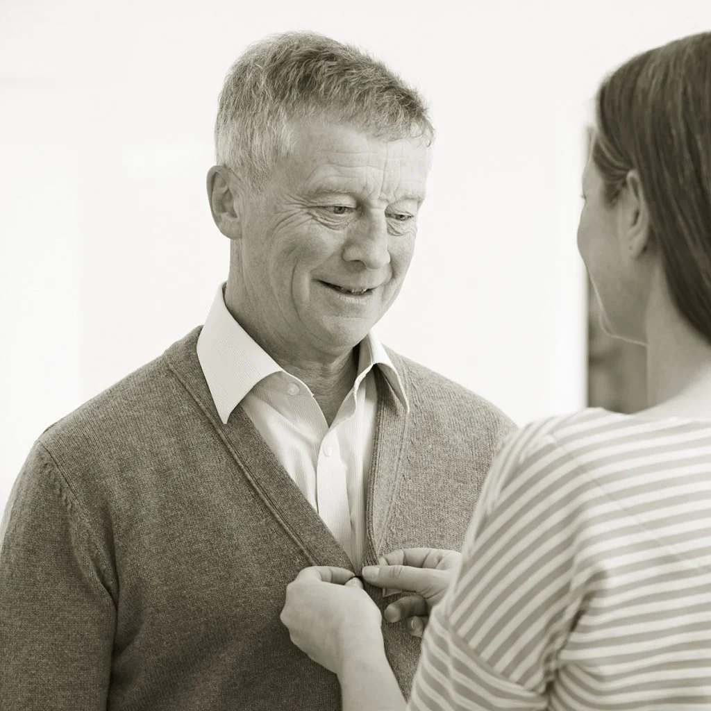 A woman pinning a boutonniere on a man in a sweater, both smiling.