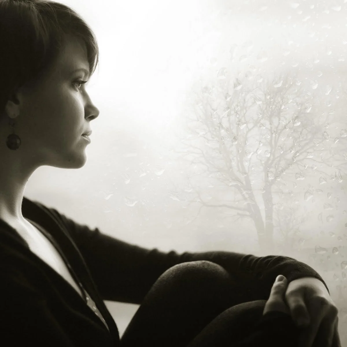 A black-and-white photo of a woman sitting by a window, looking outside at a foggy scene with a tree and raindrops on the glass.
