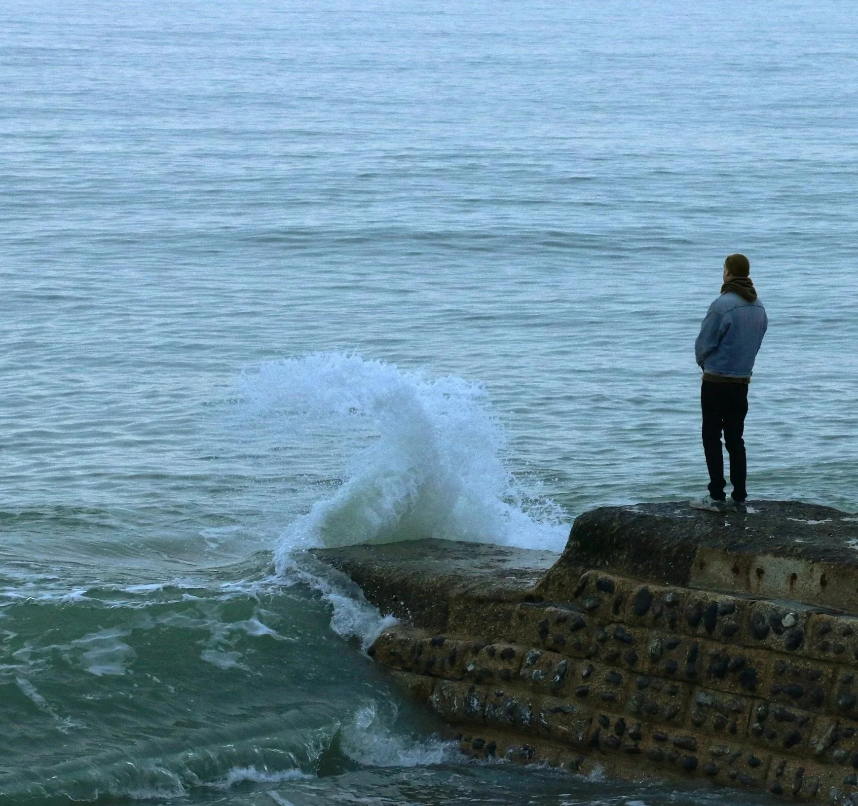 A person wearing a jacket and scarf standing on rocks by the ocean, looking at the water and waves.
