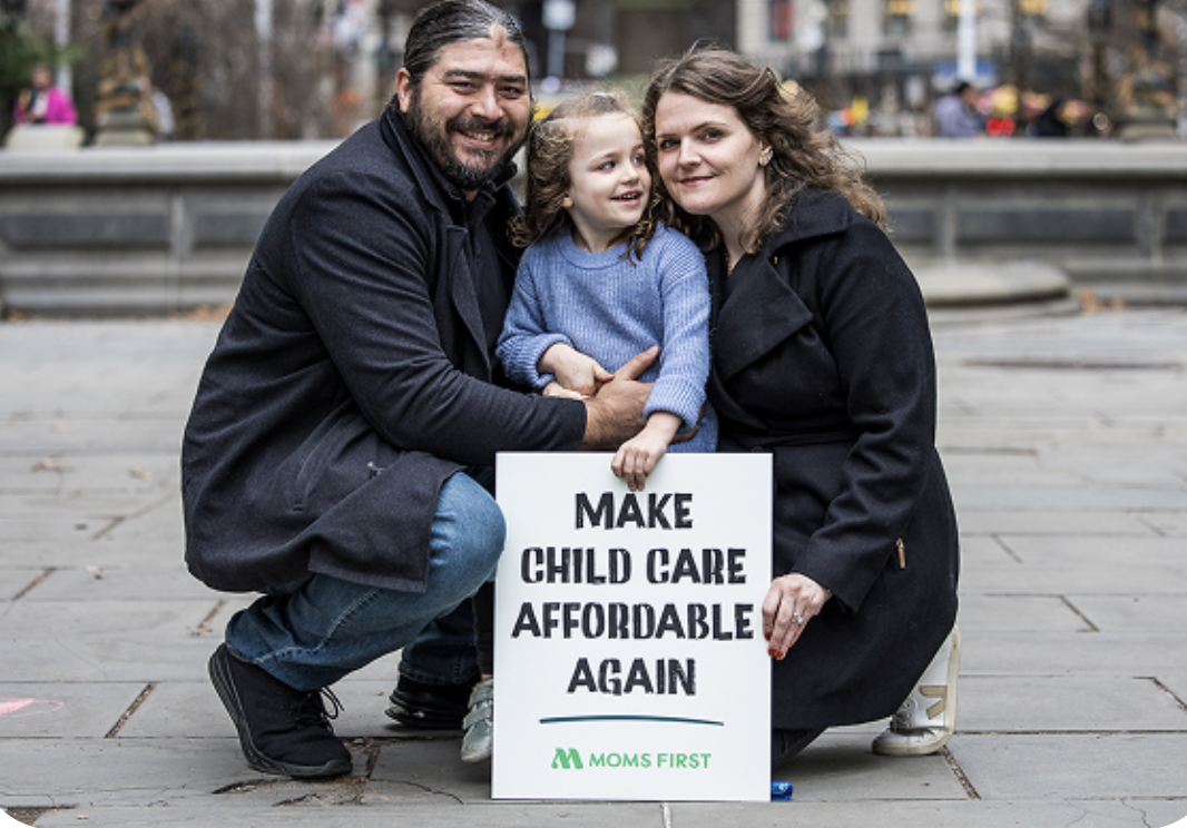 Family of three sitting on a city sidewalk, holding a sign that says 'Make Child Care Affordable Again' with 'Moms First' logo, during a protest or rally.
