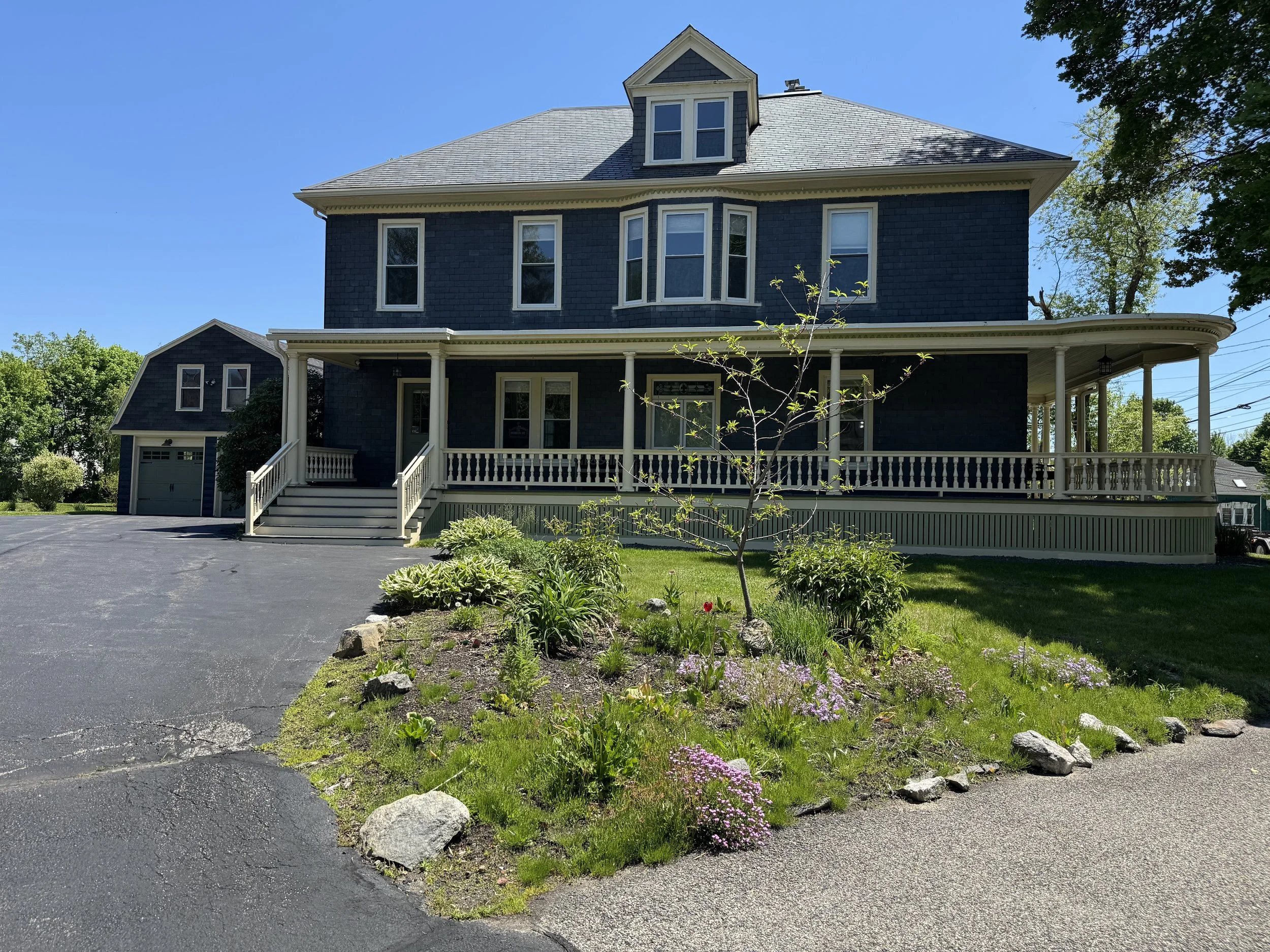 A large, dark blue, three-story house with a front porch, multiple windows, and a small dormer window on the roof. There is a driveway and a small garden with various plants and rocks in the front yard, and the sky is clear and blue.